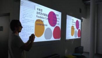 Louis Hoebregts speaking in front of two large screens with blobs shapes and Chinese characters on the screens.